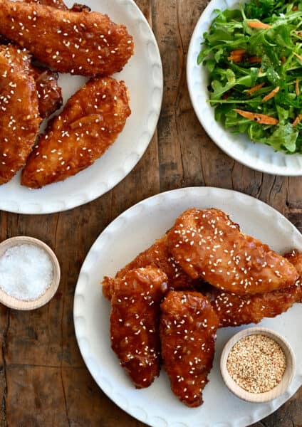 Orange chicken tenders on a plate garnished with sesame seeds next to a plate topped with mixed salad greens.