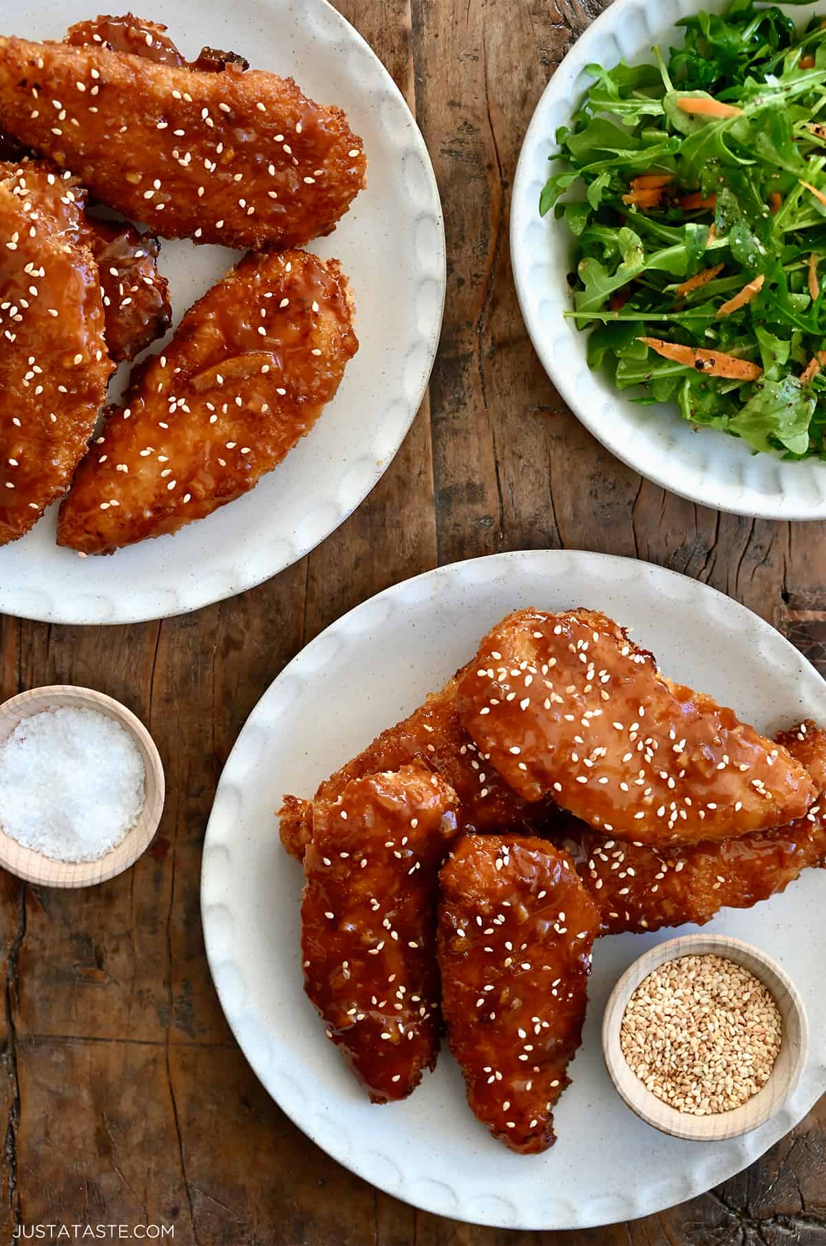 Orange chicken tenders on a plate garnished with sesame seeds next to a plate topped with mixed salad greens.