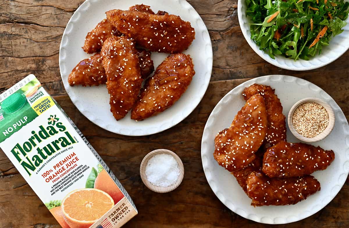 Two plates containing orange chicken tenders topped with sesame seeds next to a carton of Florida's Natural Orange Juice. 