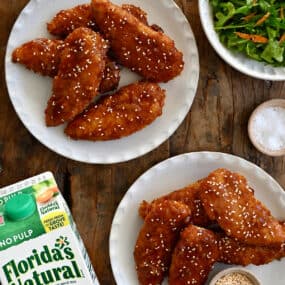A top-down view of two plates piled high with orange chicken tenders next to a carton of Florida's Natural Orange Juice.