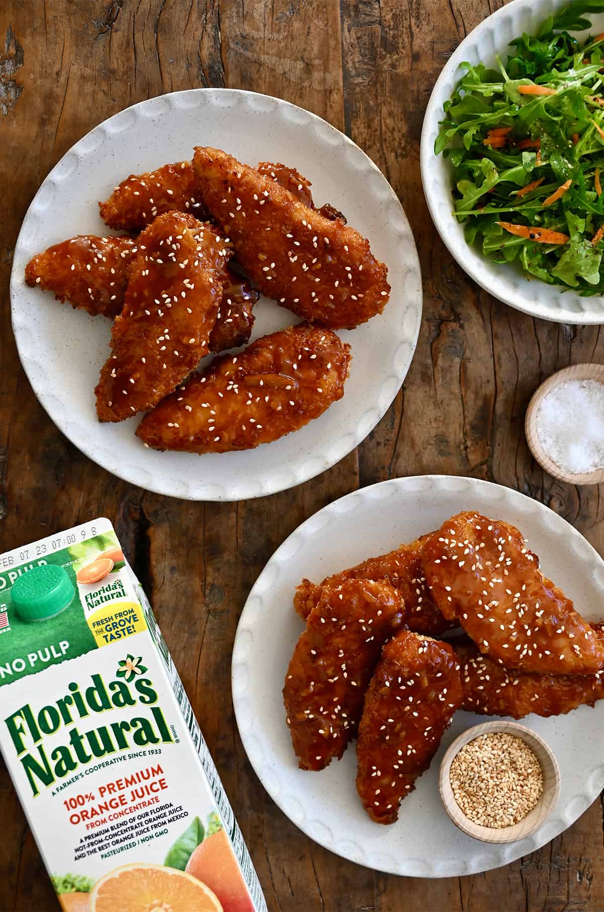 A top-down view of two plates piled high with orange chicken tenders next to a carton of Florida's Natural Orange Juice.