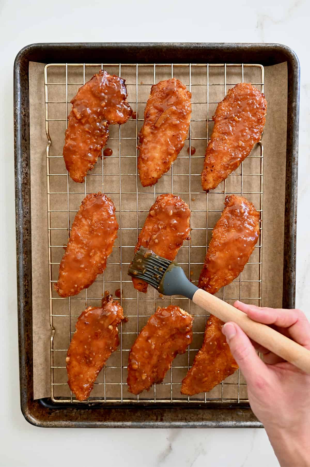 Orange sauce being brushed atop baked chicken tenders on a wire rack on a baking sheet.