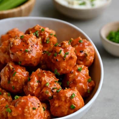 Buffalo chicken meatballs in a bowl garnished with chopped scallions near a bowl containing blue cheese dressing.