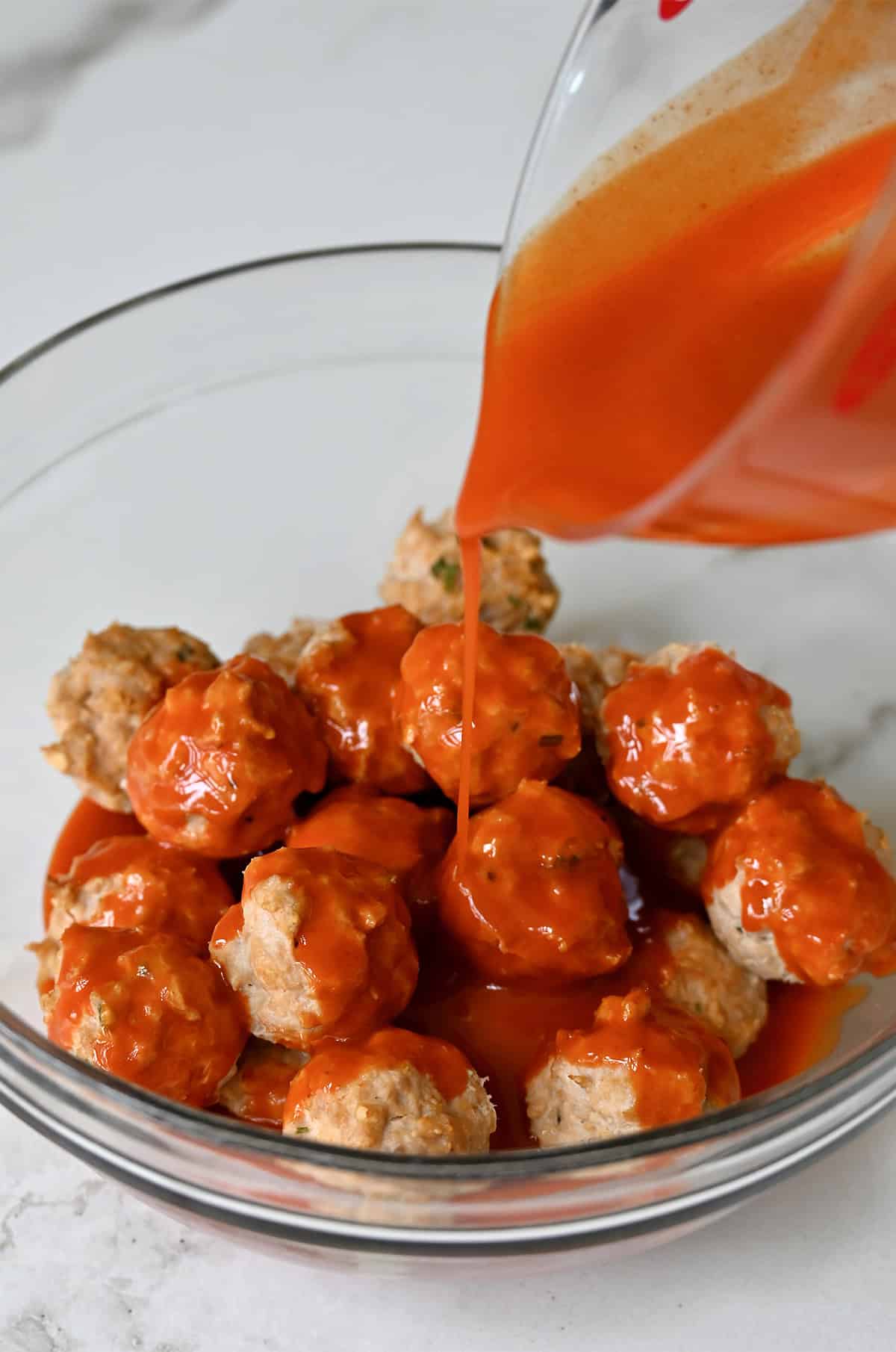 Buffalo sauce being poured over mini chicken meatballs in a bowl.