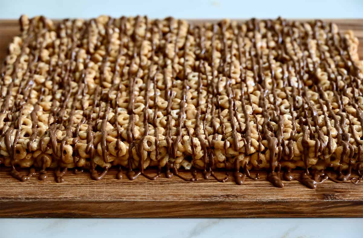 Marshmallow Cheerios bars drizzled with melted chocolate on a cutting board.
