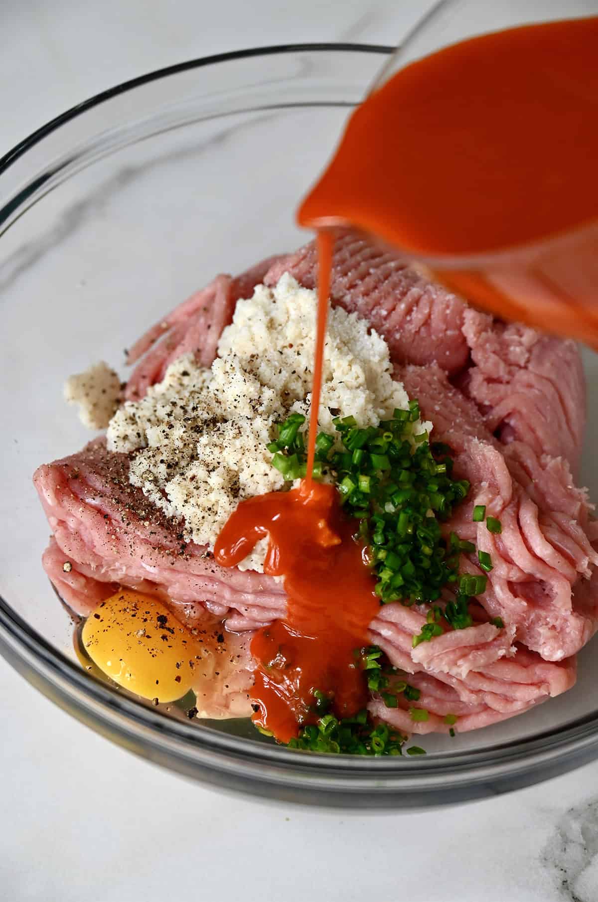 Buffalo sauce being poured over ground chicken, milk-soaked breadcrumbs, chopped chives and an egg in a large bowl.