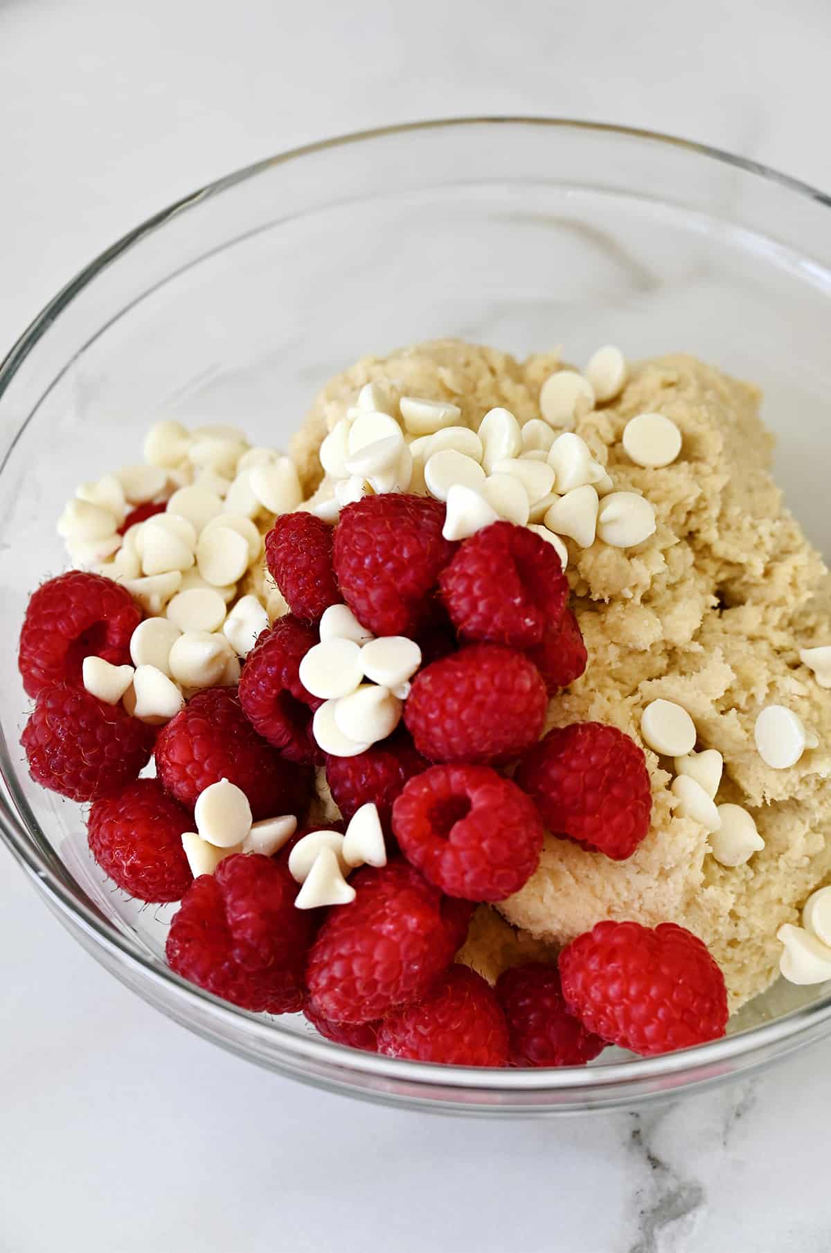 A glass bowl containing scone dough, white chocolate chips and fresh raspberries.