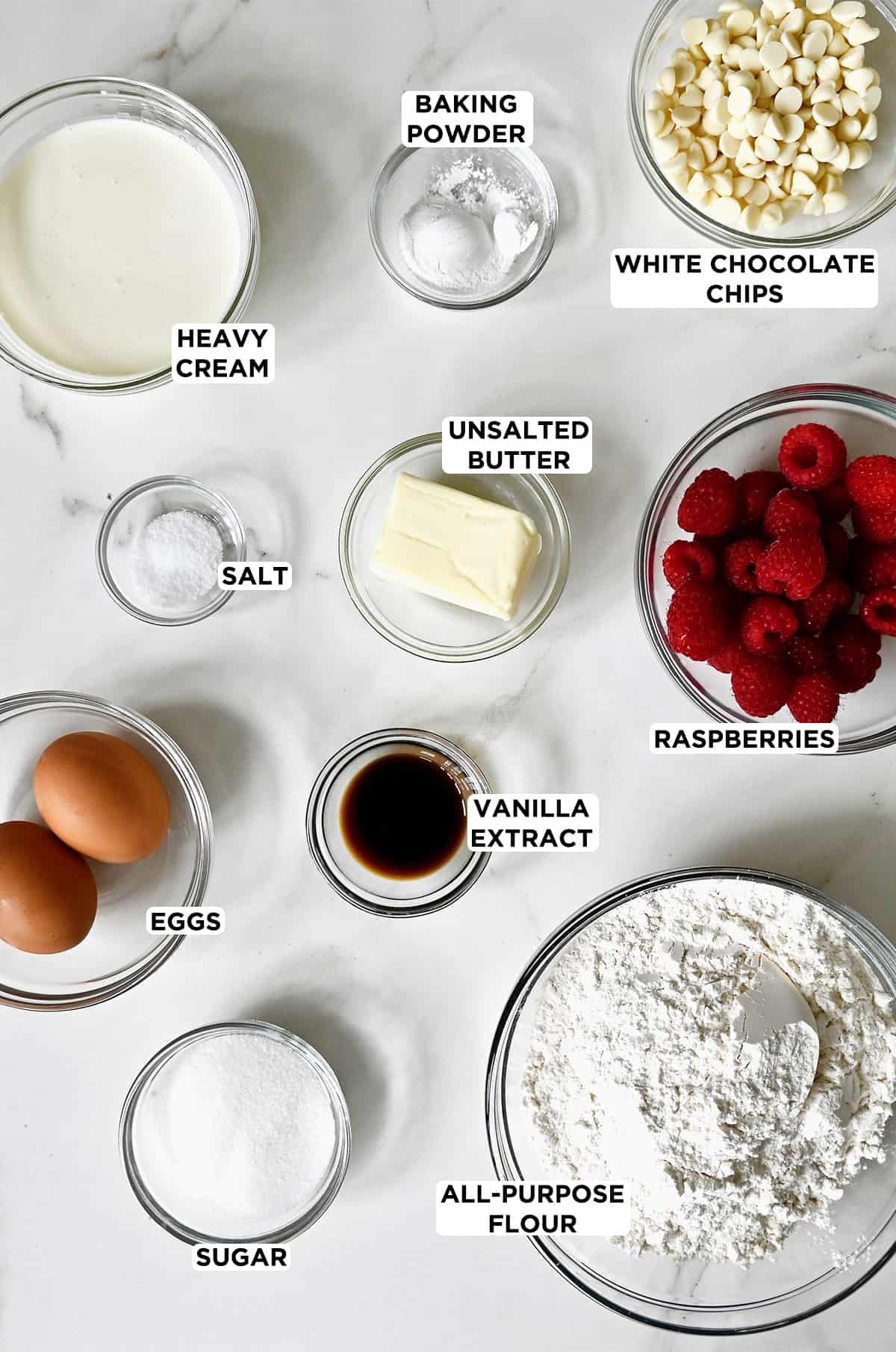 Various sizes of glass bowls containing ingredients for raspberry scones, including heavy cream, baking powder, white chocolate chips,