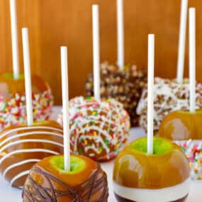 Caramel apples decorated with different toppings, including drizzled with milk chocolate, chopped walnuts, rainbow sprinkles and white chocolate.