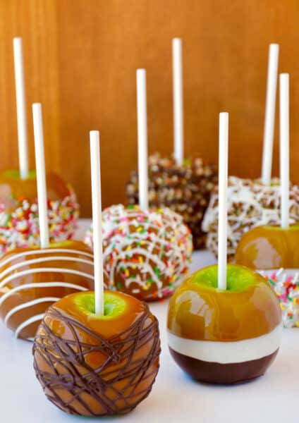 Caramel apples decorated with different toppings, including drizzled with milk chocolate, chopped walnuts, rainbow sprinkles and white chocolate.