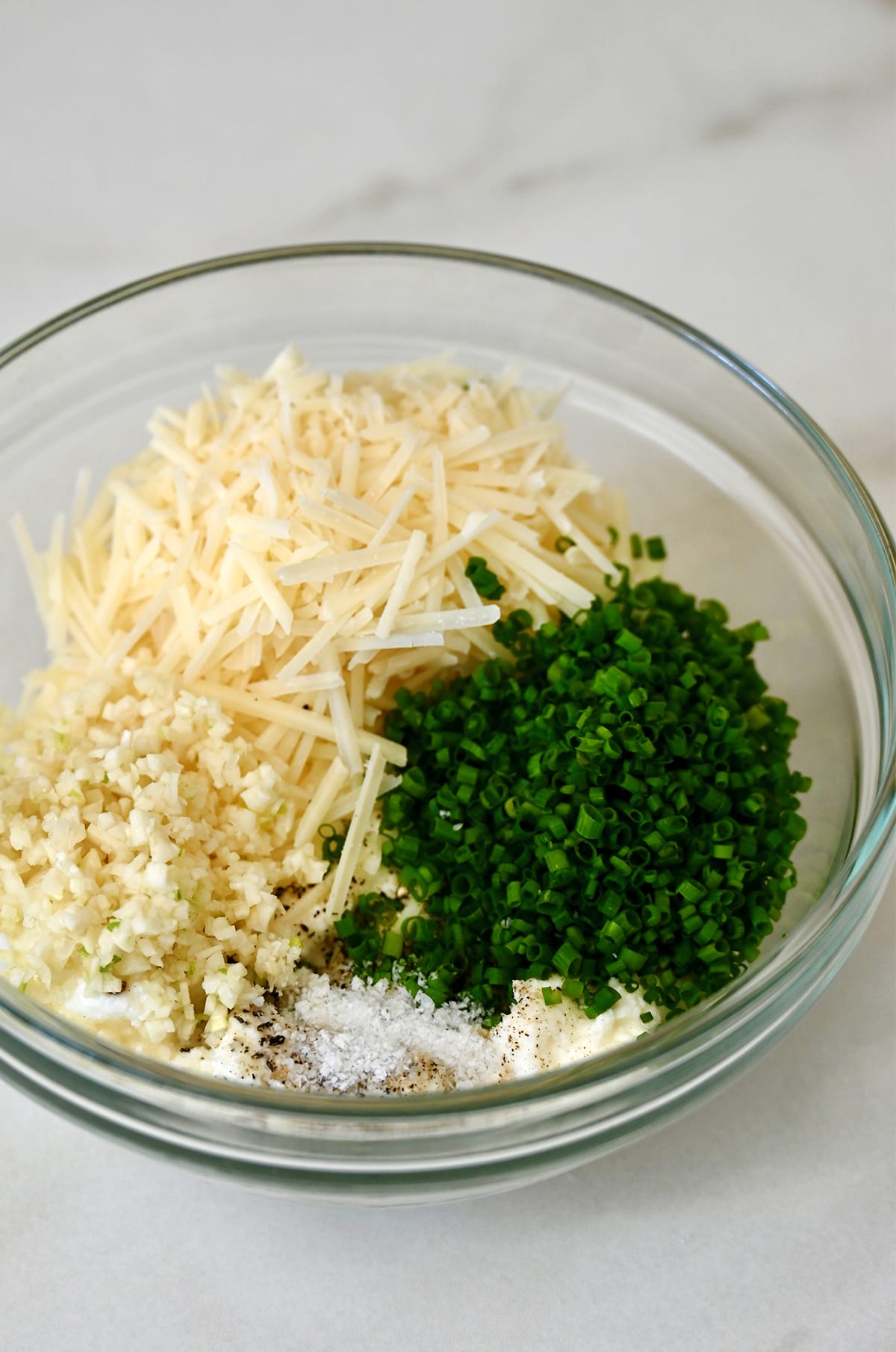 A glass bowl containing shredded Parmesan and mozzarella cheeses, chopped fresh chives, salt, pepper and shredded rotisserie chicken.