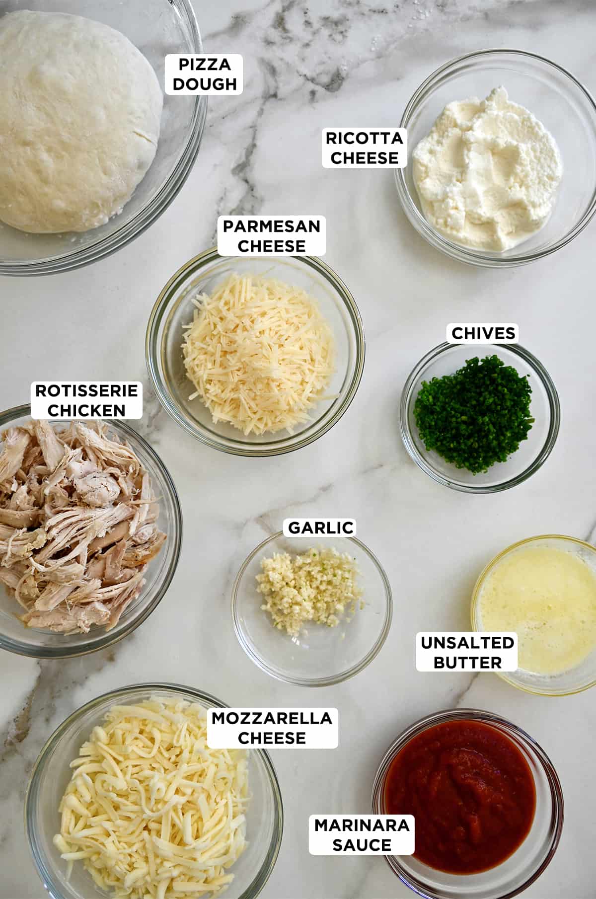 Glass bowls containing the ingredients needed to make cheesy chicken calzones, including pizza dough, ricotta cheese, chives, shredded Parmesan cheese, minced garlic, melted unsalted butter, shredded mozzarella cheese, marinara sauce and shredded rotisserie chicken. 