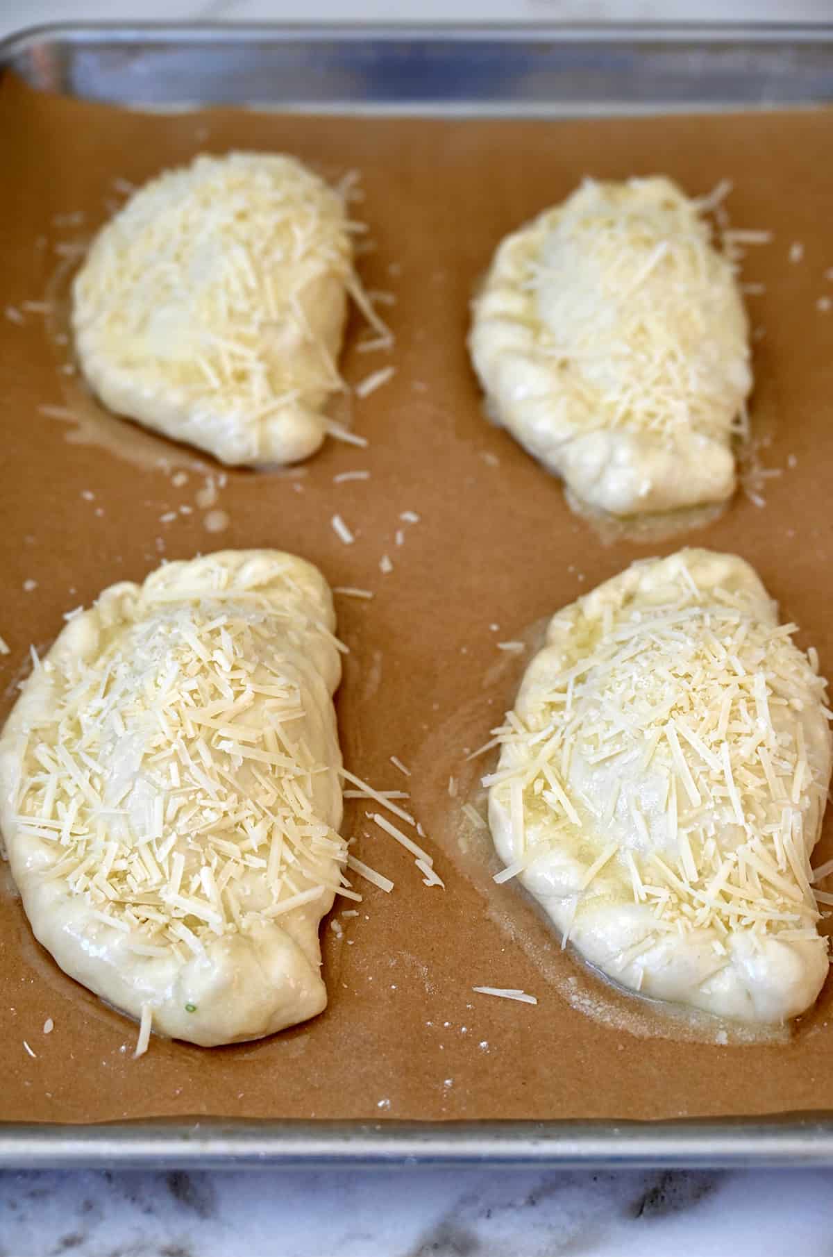 Four unbaked calzones brushed with melted butter and sprinkled with shredded Parmesan cheese on a parchment paper-lined baking sheet.