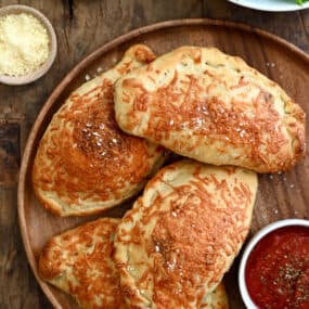 Four perfectly golden brown calzones on a dinner plate with a small bowl containing marinara sauce.