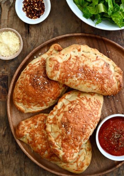 Four perfectly golden brown calzones on a dinner plate with a small bowl containing marinara sauce.