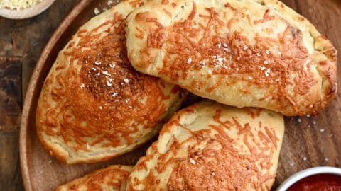 Four perfectly golden brown calzones on a dinner plate with a small bowl containing marinara sauce.