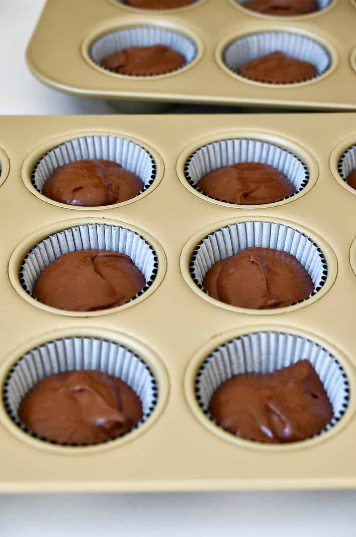 Two muffin tins with cupcake liners filled with chocolate cake batter.