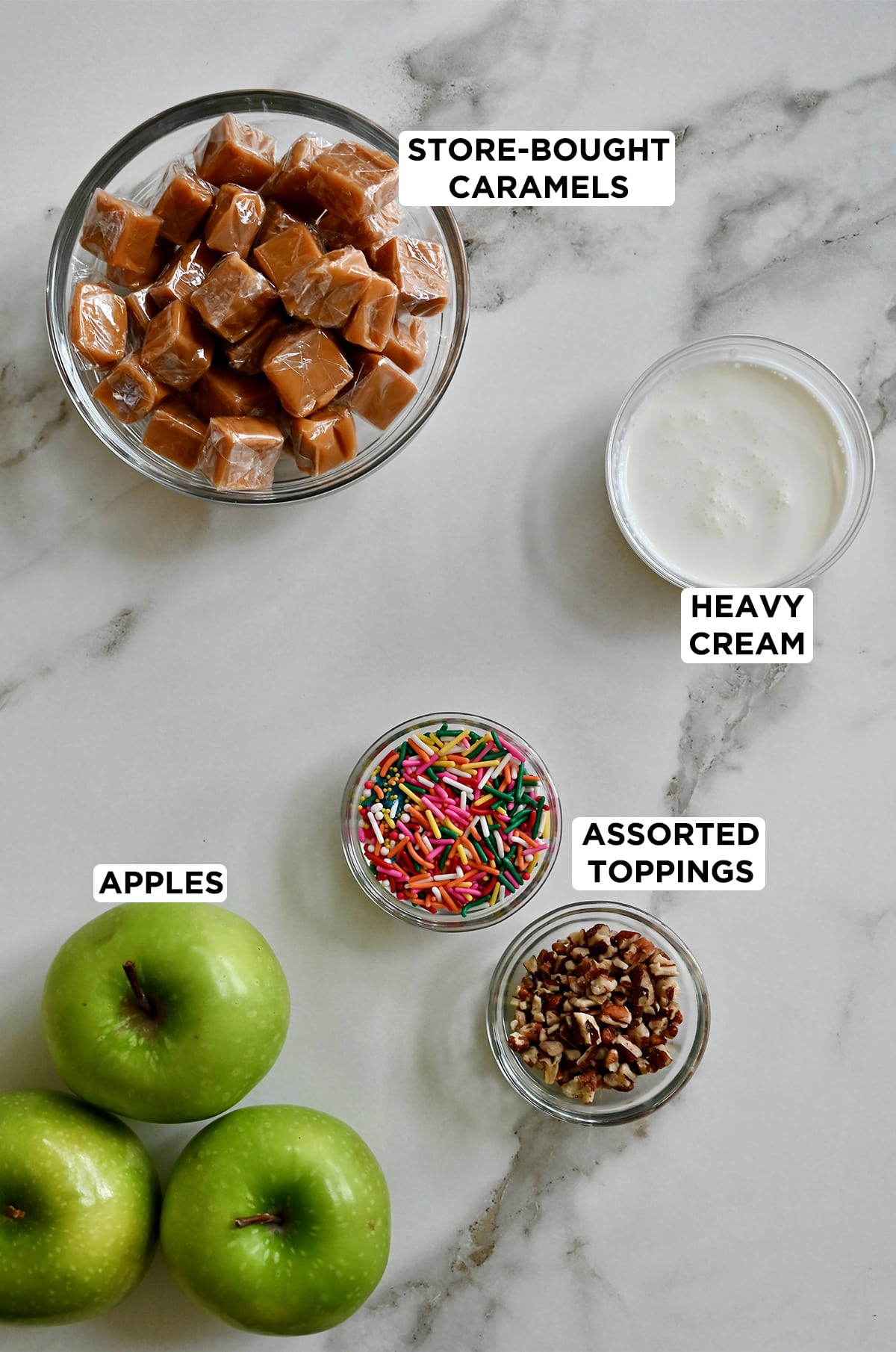 Granny Smith apples next to three bowls containing soft caramels, heavy cream, rainbow sprinkles and chopped pecans. 