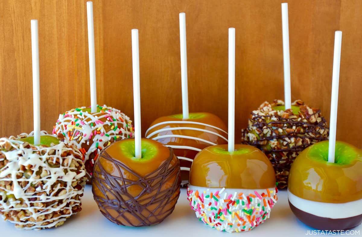 Two rows of homemade caramel apples with various toppings, including chopped nuts, white chocolate and rainbow sprinkles, milk chocolate drizzle, and dried fruit.