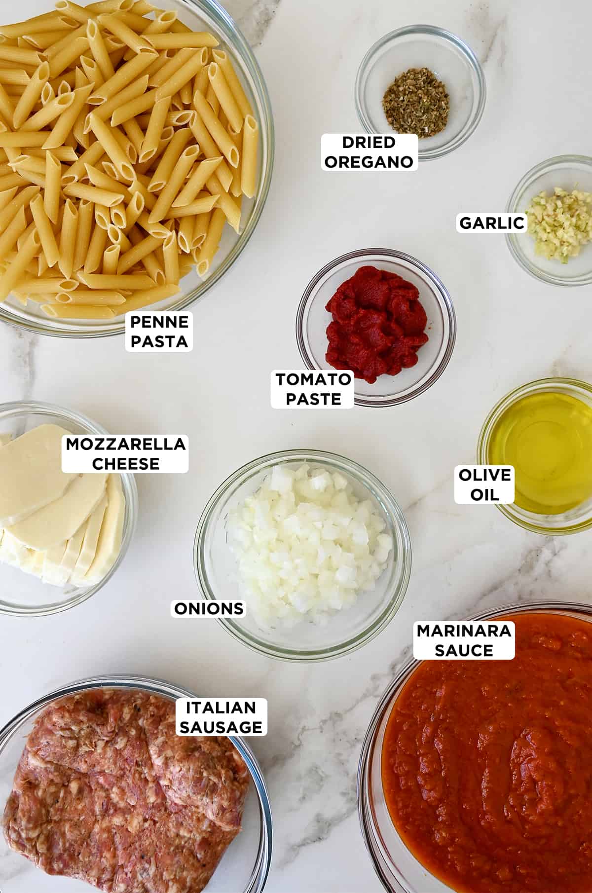 All the ingredients needed to make baked penne in clear glass bowls, including uncooked penne, dried oregano, minced garlic, tomato paste, olive oil, diced onions, marinara sauce, Italian sausage and slices of fresh mozzarella cheese.