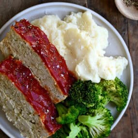 Two slices of classic chicken meatloaf on a dinner plate with steamed broccoli and mashed potatoes.