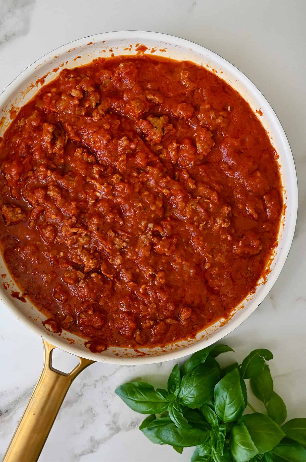 A large skillet containing sausage bolognese sauce next to fresh basil leaves.