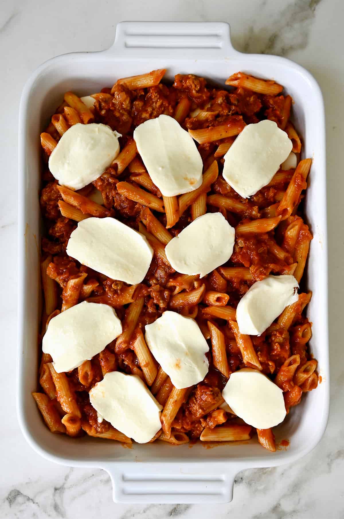 Slices of fresh mozzarella atop penne pasta with a bolognese sauce in a white baking dish.