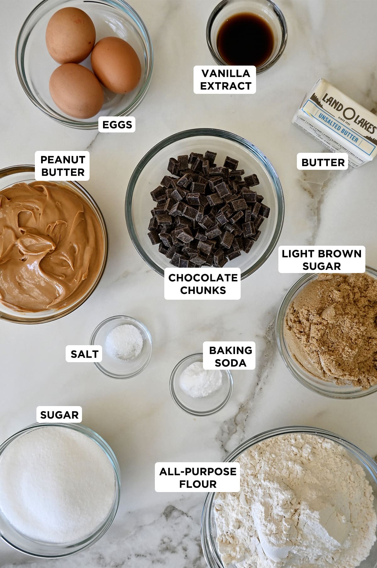 Various sizes of clear bowls containing the ingredients needed to make peanut butter chocolate chip cookies: eggs, vanilla extract, butter, chocolate chips, light brown sugar, baking soda, salt, flour, sugar and creamy peanut butter.