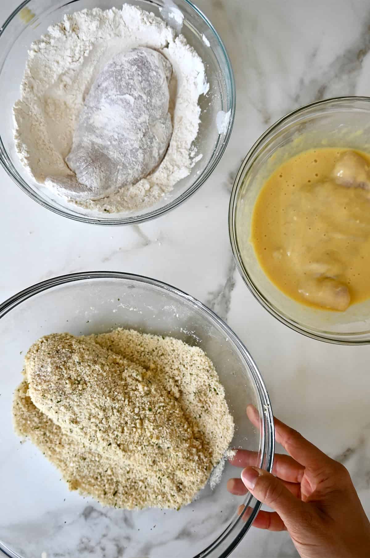 Three glass bowls for dredging chicken breasts; one contains panko breadcrumbs, another has an egg wash with Dijon mustard, and the third has flour.