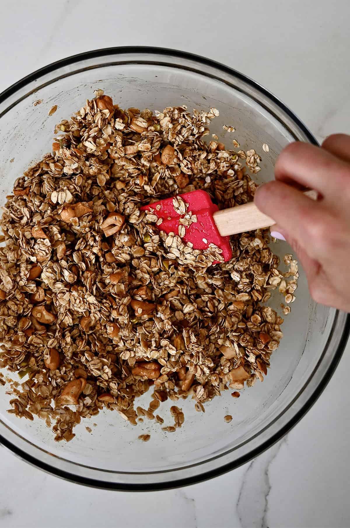 Toasted oats and nuts are being stirred with a mixture of oil, honey, vanilla and cinnamon in a large mixing bowl.
