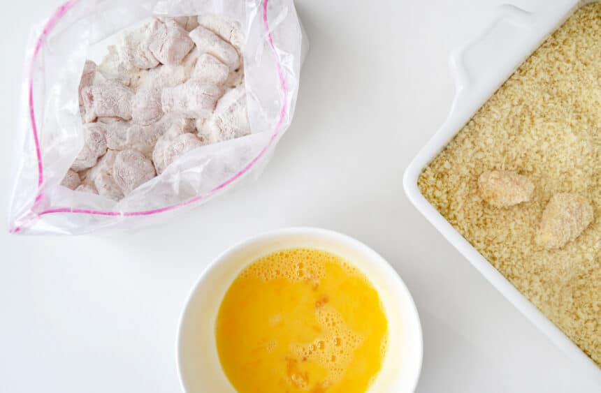 A top-down view of a plastic bag containing chicken nuggets and flour next to a bowl containing an egg wash and a shallow dish containing breadcrumbs