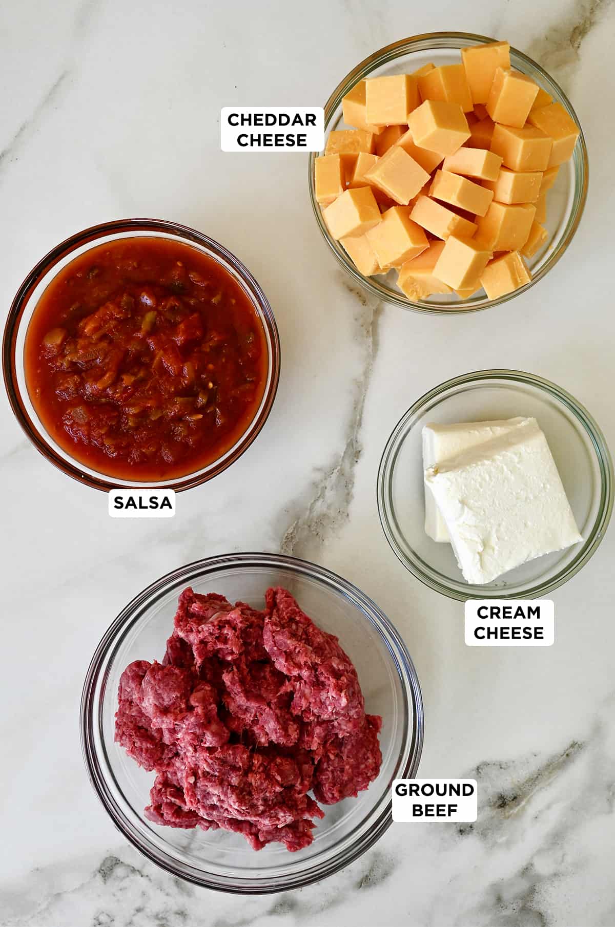 Four glass bowls containing ground beef, salsa, cream cheese and cubed cheddar cheese.