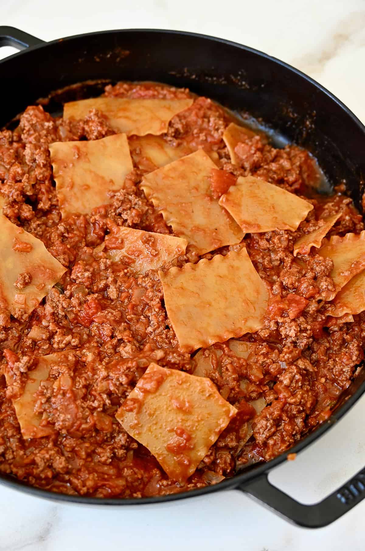 Pieces of lasagna noodles mixed into bolognese sauce in a large skillet.