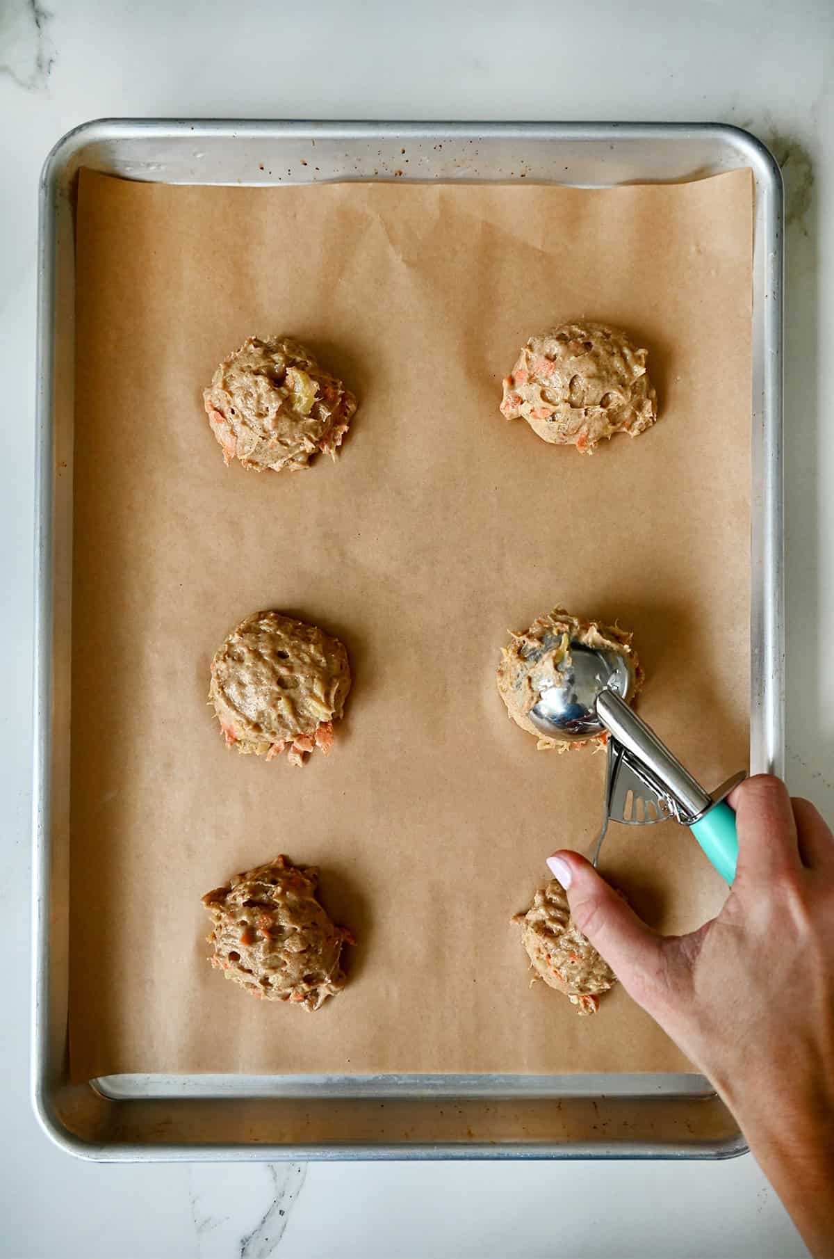 Scooping carrot cake cookie dough onto a parchment paper-lined baking sheet with an ice cream scoop.