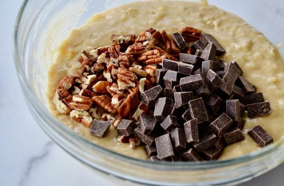 A glass bowl with banana bread batter, chopped nuts and chocolate chunks