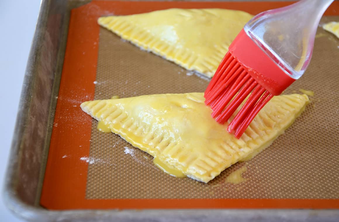 A pastry brush applies an egg wash atop unbaked pastries on a lined baking sheet