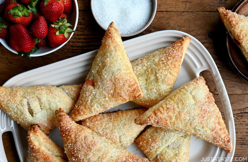 A top-down view of rhubarb turnovers on a white serving platter next to a bowl filled with fresh strawberries