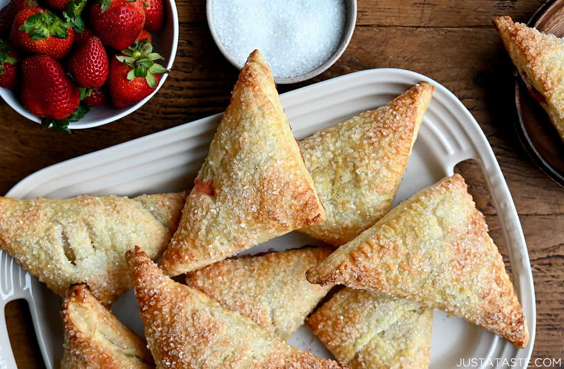A top-down view of rhubarb turnovers on a white serving platter next to a bowl filled with fresh strawberries