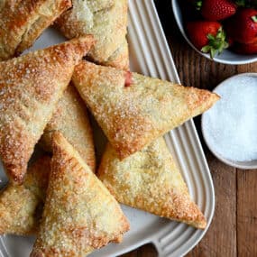 Strawberry Rhubarb Turnovers on a white serving plate next to a bowl containing fresh strawberries and a bowl containing sanding sugar