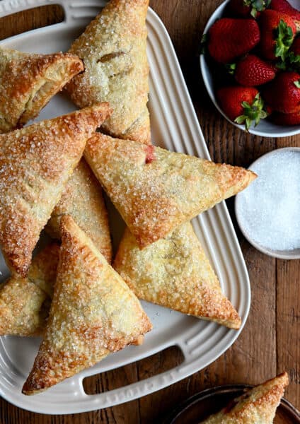 Strawberry Rhubarb Turnovers on a white serving plate next to a bowl containing fresh strawberries and a bowl containing sanding sugar
