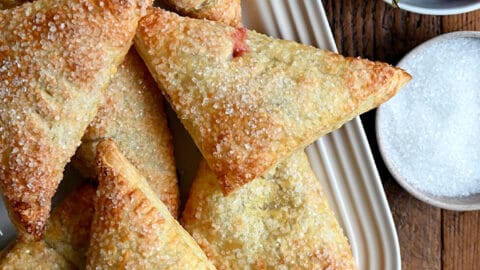 Strawberry Rhubarb Turnovers on a white serving plate next to a bowl containing fresh strawberries and a bowl containing sanding sugar