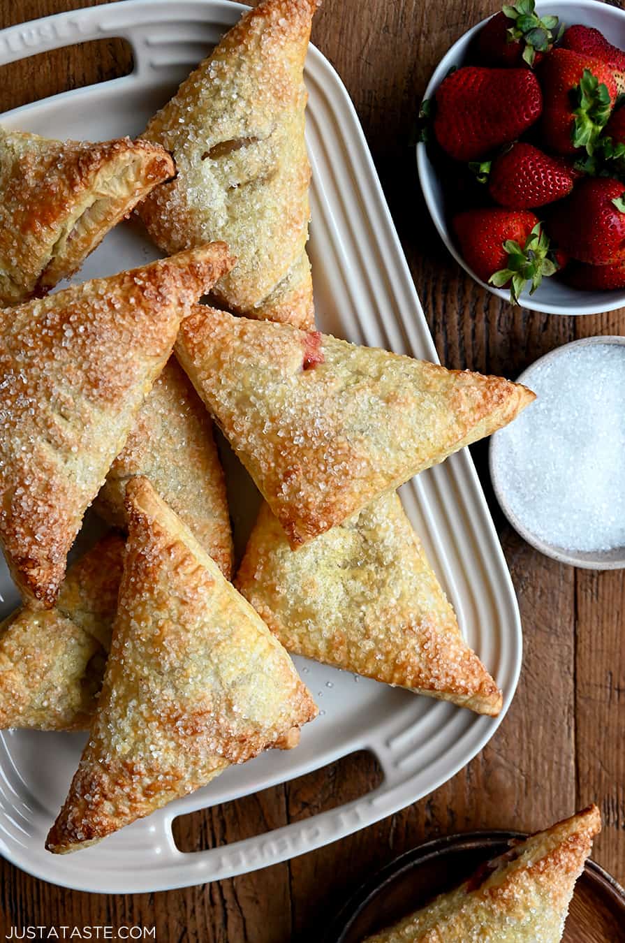 Strawberry Rhubarb Turnovers on a white serving plate next to a bowl containing fresh strawberries and a bowl containing sanding sugar