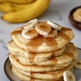 A tall stack of fluffy banana pancakes topped with slices of banana and warm maple syrup.