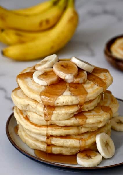 A tall stack of fluffy banana pancakes topped with slices of banana and warm maple syrup.