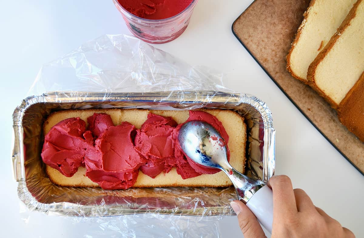 A hand holding an ice cream scooper places mounds of raspberry sorbet atop pound cake in an aluminum tray.