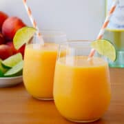 Two wine glasses filled with Peach Wine Slushies garnished with a lime wedge and an orange striped straw. A bottle of white wine and a bowl of peaches are in the background.