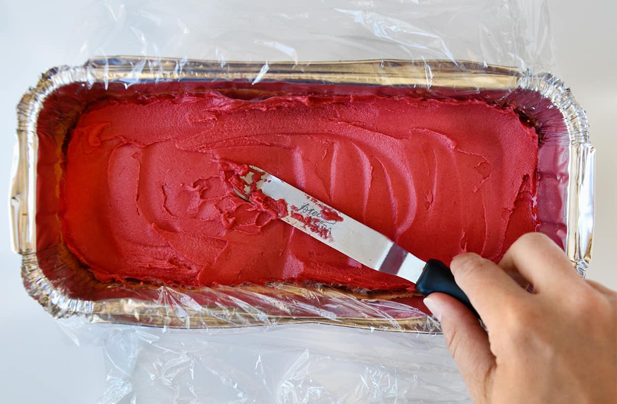 A hand holding an offset spatula spreads raspberry sorbet over pound cake in a plastic wrap-lined aluminum tray.