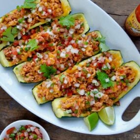 A white plate of zucchini boats surrounded by toppings and hot sauce