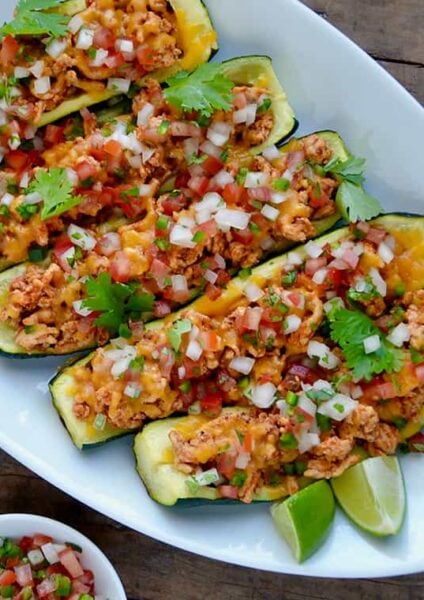 A white plate of zucchini boats surrounded by toppings and hot sauce