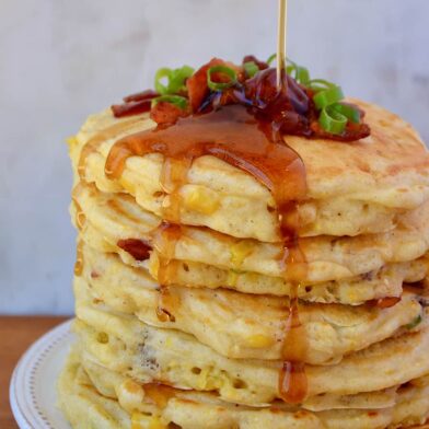 Tall stack of Sweet Corn Pancakes with Bacon topped with sliced scallions and drizzled with maple syrup